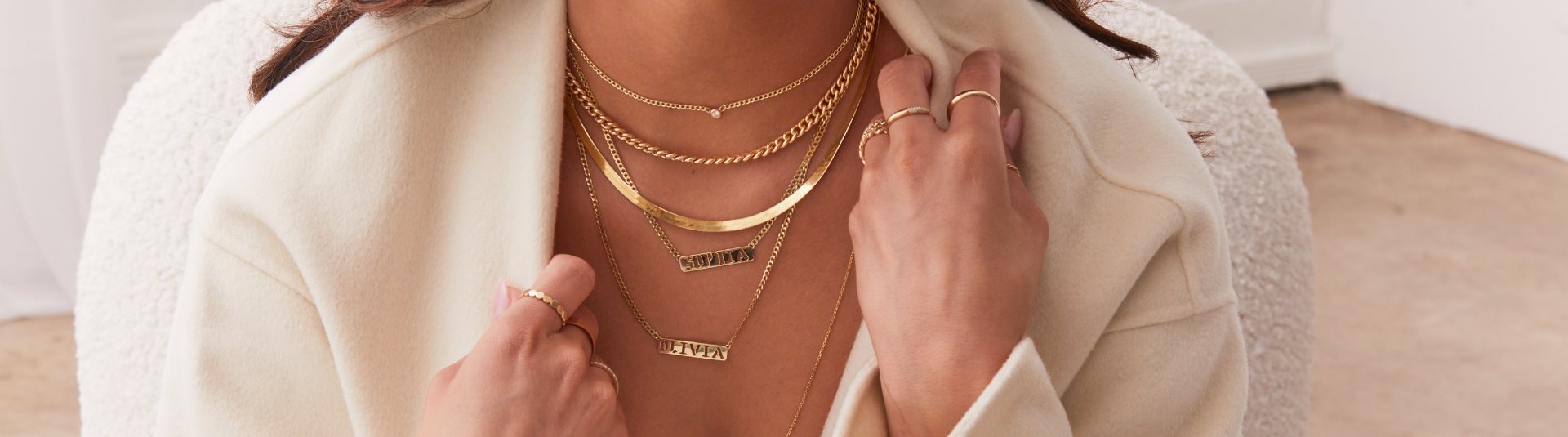 Close up of woman's neck with layered 14K solid gold chains - herringbone, diamond bezel, chunky chains, personalized name necklaces. Wearing white cardigan, sitting in white chair with hands holding her cardigan and 14K solid gold rings on.