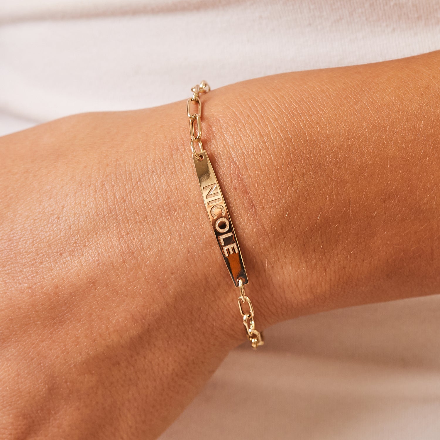 Lifestyle photo closeup on model's wrist with 14K yellow gold bracelet with oval link chain and solid name plate with engraved name on it and lobster clasp.