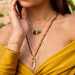 A close-up lifestyle shot of a model wearing a layered gold necklace set with a white tooth pendant, a green bead accent, and brown beaded strands, paired with a mustard yellow top.