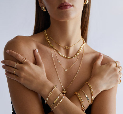 Woman wearing 14K solid  yellow gold hoop earrings, stacked ID, cuban link, and paperclip chain bracelets, and layered necklaces with natural diamonds and personalized name jewelry.