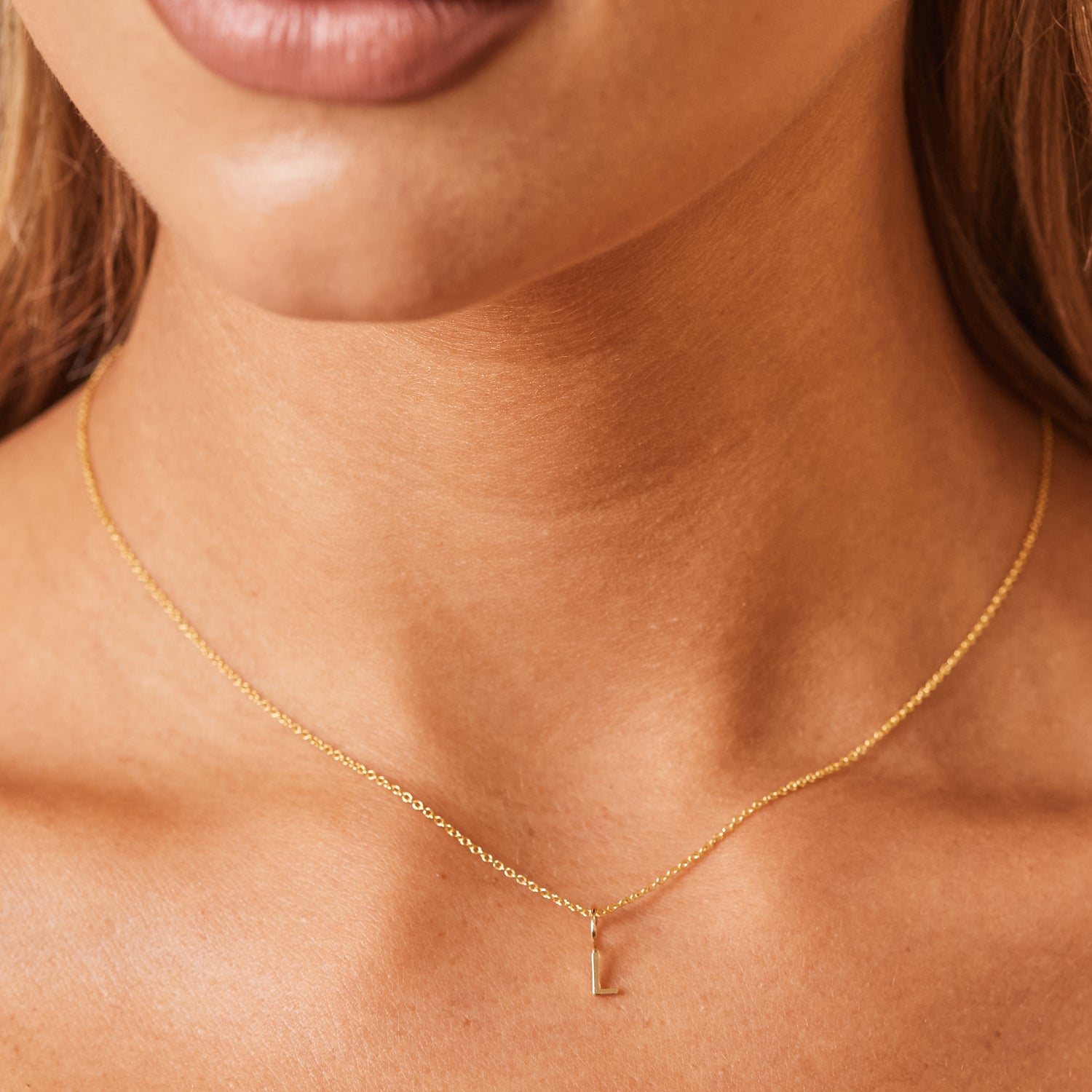 Close up model shot of 14K yellow gold initial charm in print, block font on a 5mm round bail and hanging from a dainty cable chain. 16 inch chain falls slightly below collarbone.