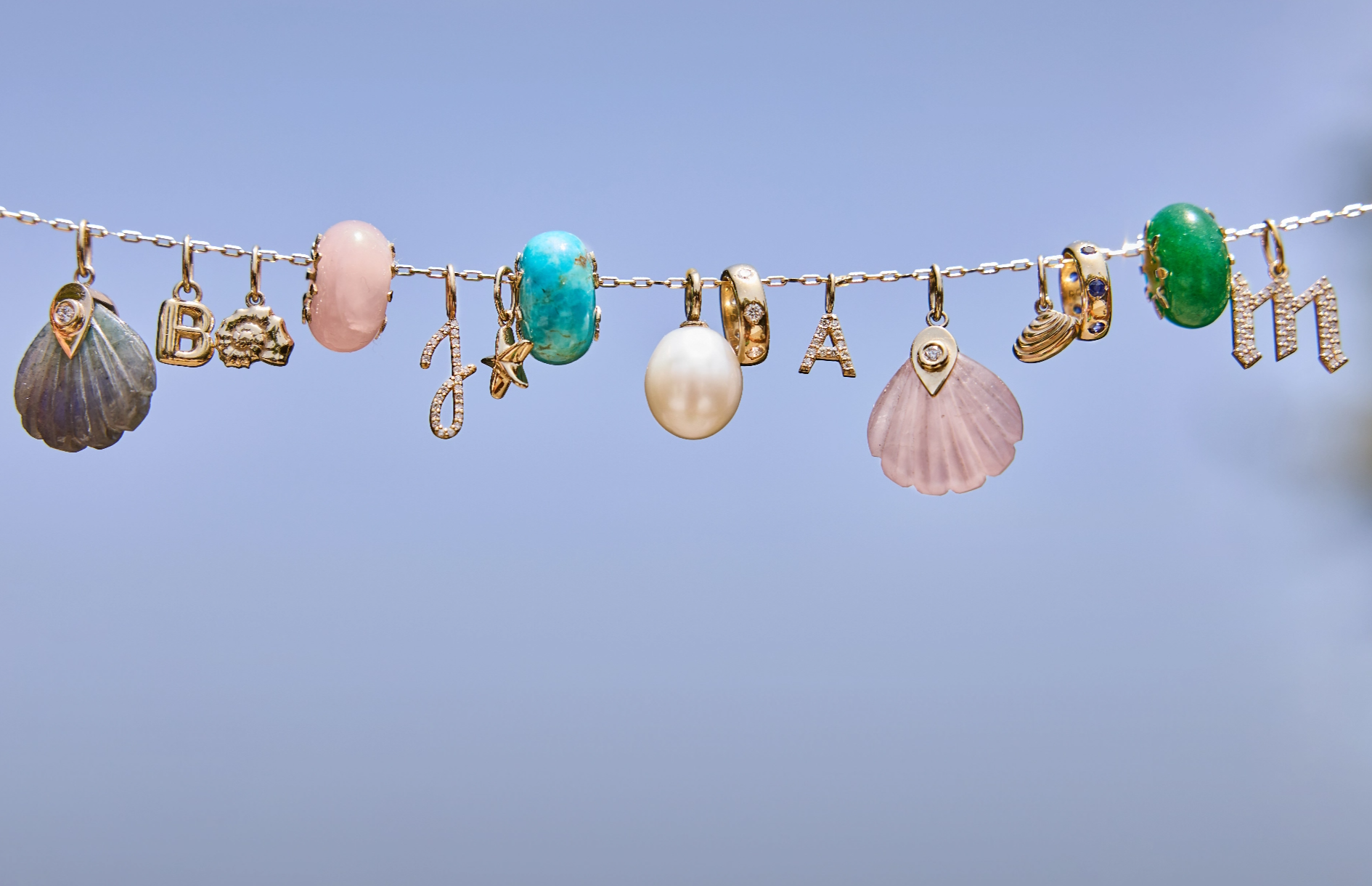 A row of charms on a 14k soid gold chain with slider charms, gemstone charms, initial charms, natural diamond charms, seashell charms.