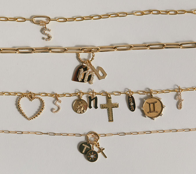 Flat lay of paperclip chains in 4 different sizes with 14k solid gold charm connectors, initial charms in different fonts, heart charm, cross charms, pave charms, zodiac charm.