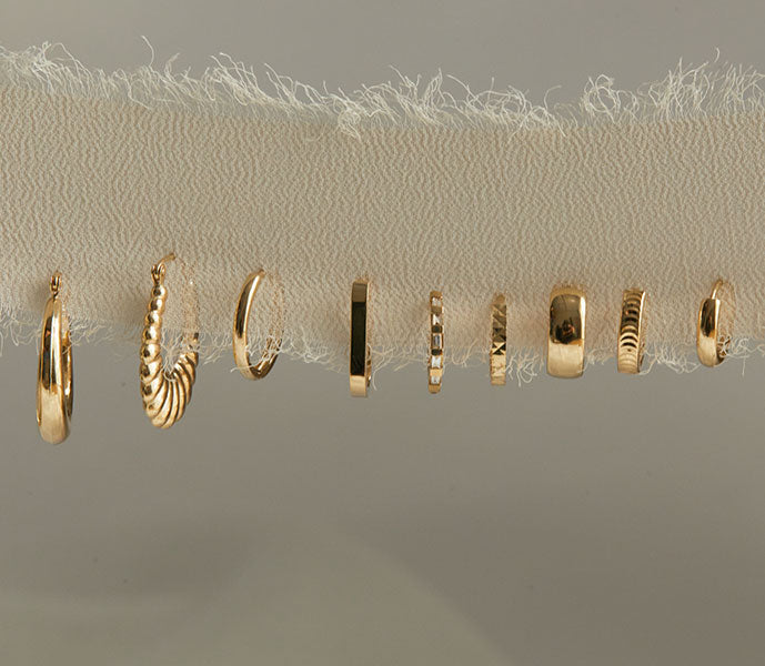 A row of 14K solid gold hoop earrings and huggie earrings in solid, diamond, and croissant textures.