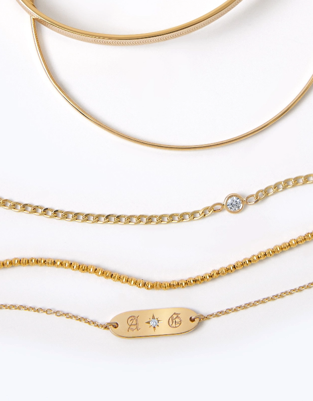 Flat lay of 14K solid gold bangles, cuban link bracelet with solitaire bezel diamond, disco ball chain, engraved charm with diamond starset.