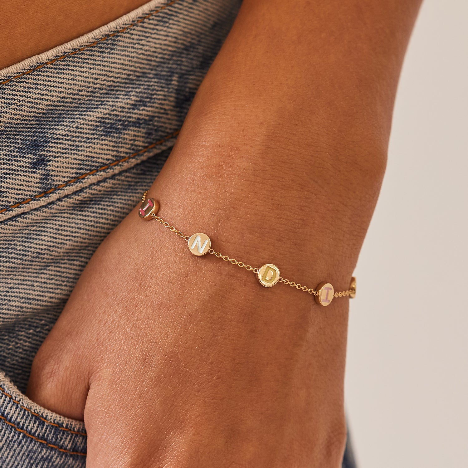 Close up of model's wrist with custom 14K yellow gold bracelet with cable chain, lobster extender clasp, and 5 gold discs with letters in all different enamel colors. Spelling the name India (from left to right) hot pink I, light blue N, green D, pink I, blue A. All capital print font letters.