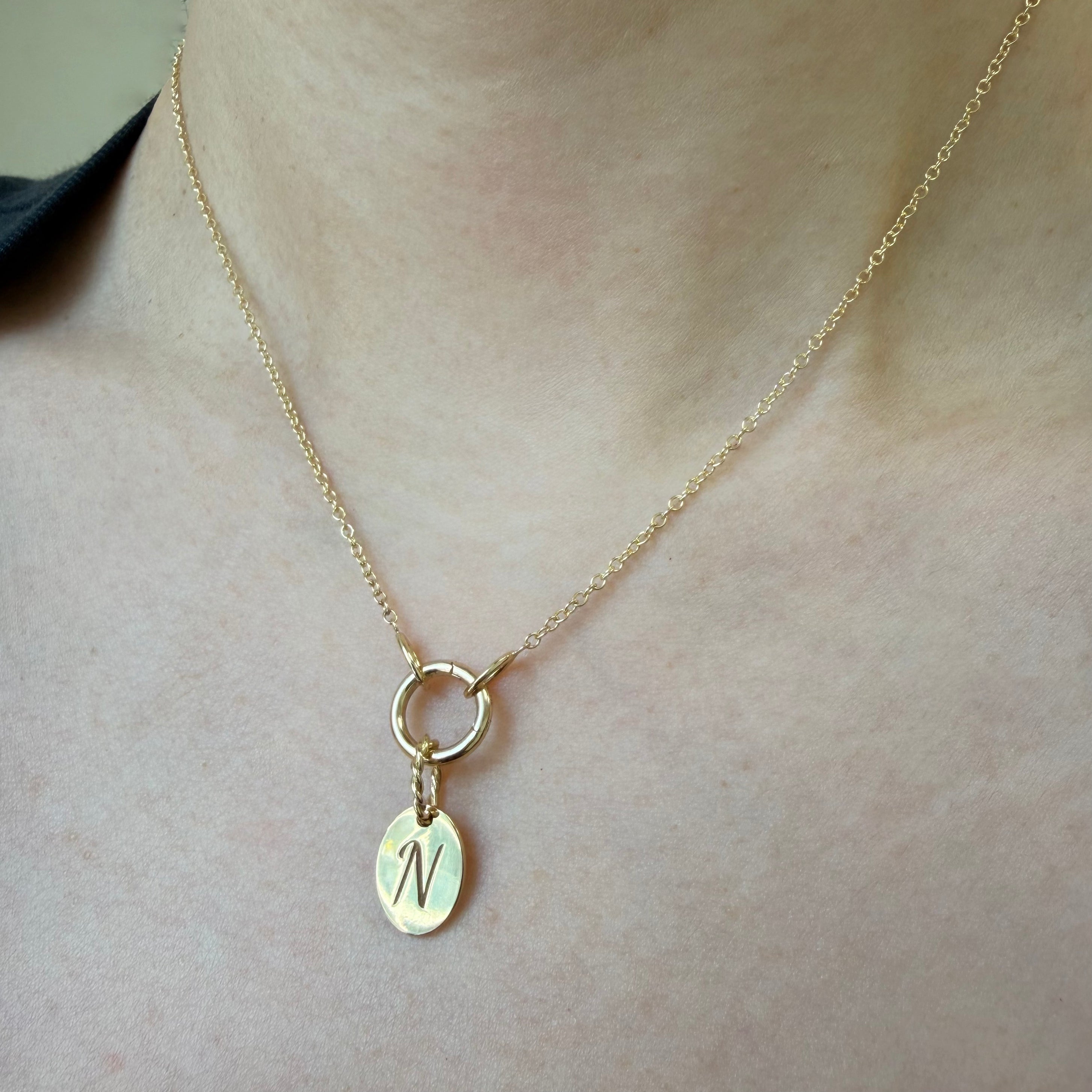 Lifestyle photo of close up on model's neck with 14K solid yellow gold disc with an engraved capital letter in script font and large bail made of twisted gold wire hanging on a charm connector necklace chain