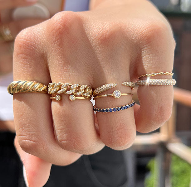 Model's hand with 14K solid gold rings on each finger. Croissant chunky ring, diamond solitaire ring, cuban link ring, dainty twisted ring, sapphire eternity band, diamond row rings, art deco rings, statement rings.