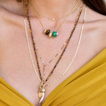 A close-up of a person wearing layered gold necklaces, including beaded chains, a small charm necklace, and a longer necklace with a white curved pendant, paired with a mustard yellow top.