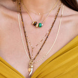 A close-up of a person wearing layered gold necklaces, including beaded chains, a small charm necklace, and a longer necklace with a white curved pendant, paired with a mustard yellow top.